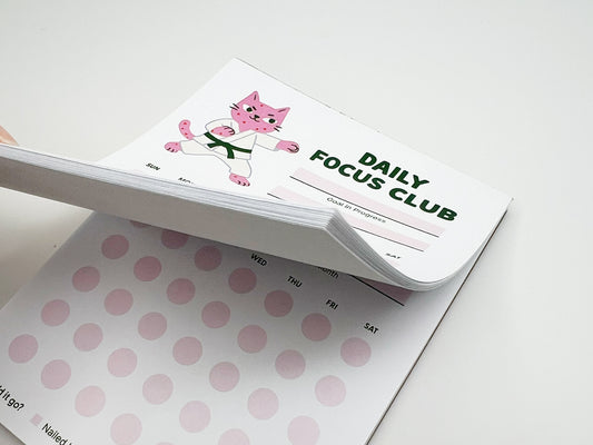 Stacked tear-off pages of Daily Focus Club monthly habit tracker notepad showing pink karate cat illustration, goal-in-progress section, and soft pink daily tracking dots labeled by day of week. Cute productivity stationery for goal setting, habit tracking, ADHD-friendly planning, and desk organization.
