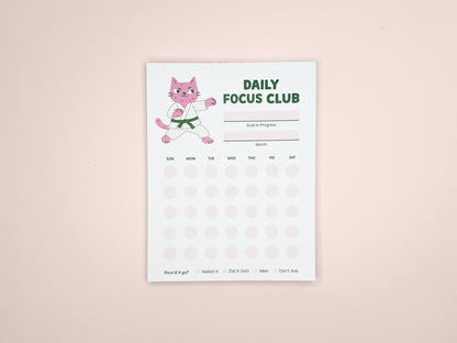 Daily Focus Club productivity notepad showing pink karate cat character, monthly habit tracker layout, goal and month sections, and soft pink dot grid. Compact desk notepad designed for gentle accountability, goal setting, and daily planning.