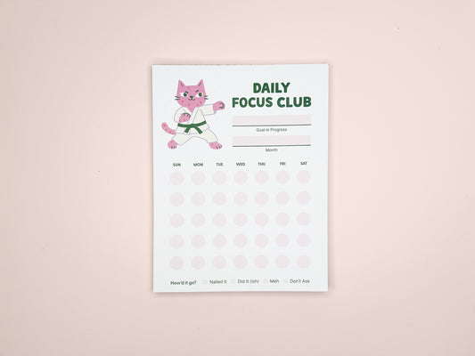 Daily Focus Club productivity notepad showing pink karate cat character, monthly habit tracker layout, goal and month sections, and soft pink dot grid. Compact desk notepad designed for gentle accountability, goal setting, and daily planning.