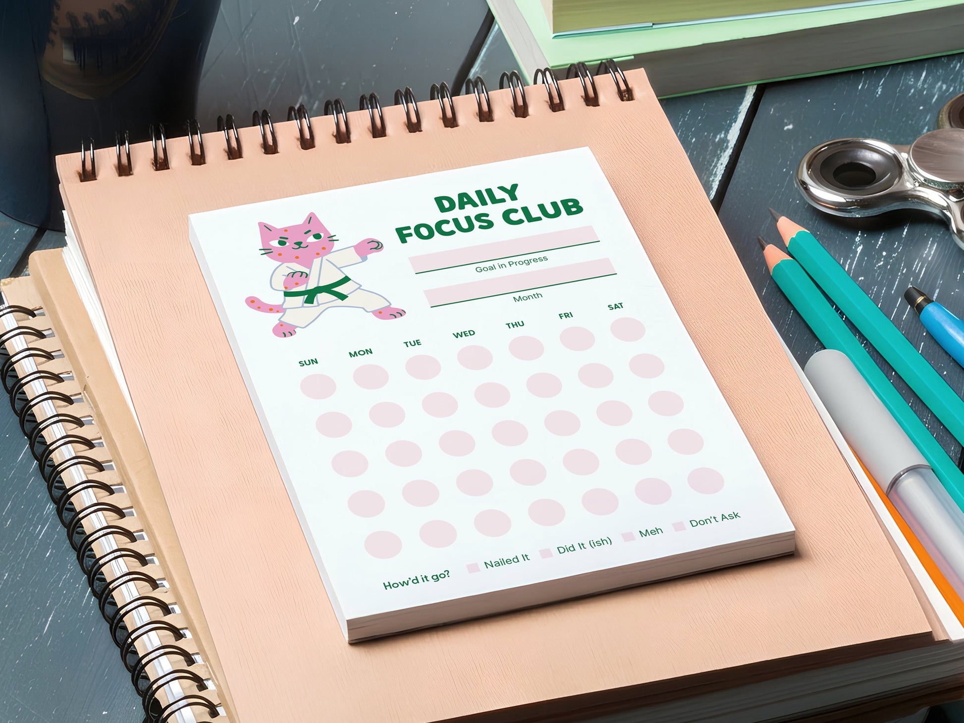 Daily Focus Club 4.25 x 5.5 inch monthly habit tracker notepad with pink karate cat illustration displayed on a peach spiral notebook beside colorful pens on a desk. Productivity notepad features goal and month sections with soft pink daily tracking dots labeled Sunday through Saturday. Cute desk stationery for goal setting, habit tracking, ADHD-friendly planning, and gentle accountability