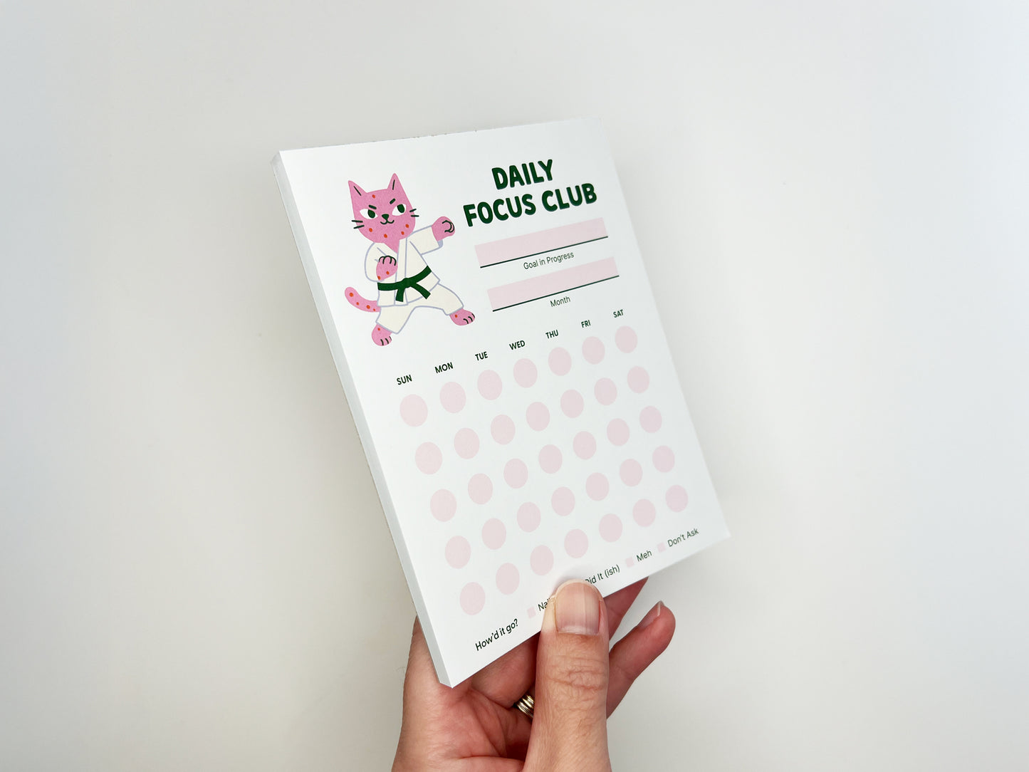 Hand holding Daily Focus Club 4.25 x 5.5 inch monthly habit tracker notepad featuring pink karate cat illustration, goal and month sections, and soft pink daily tracking dots. Cute productivity notepad for goal setting, habit tracking, ADHD-friendly planning, and desk organization by Sungy Side Up.