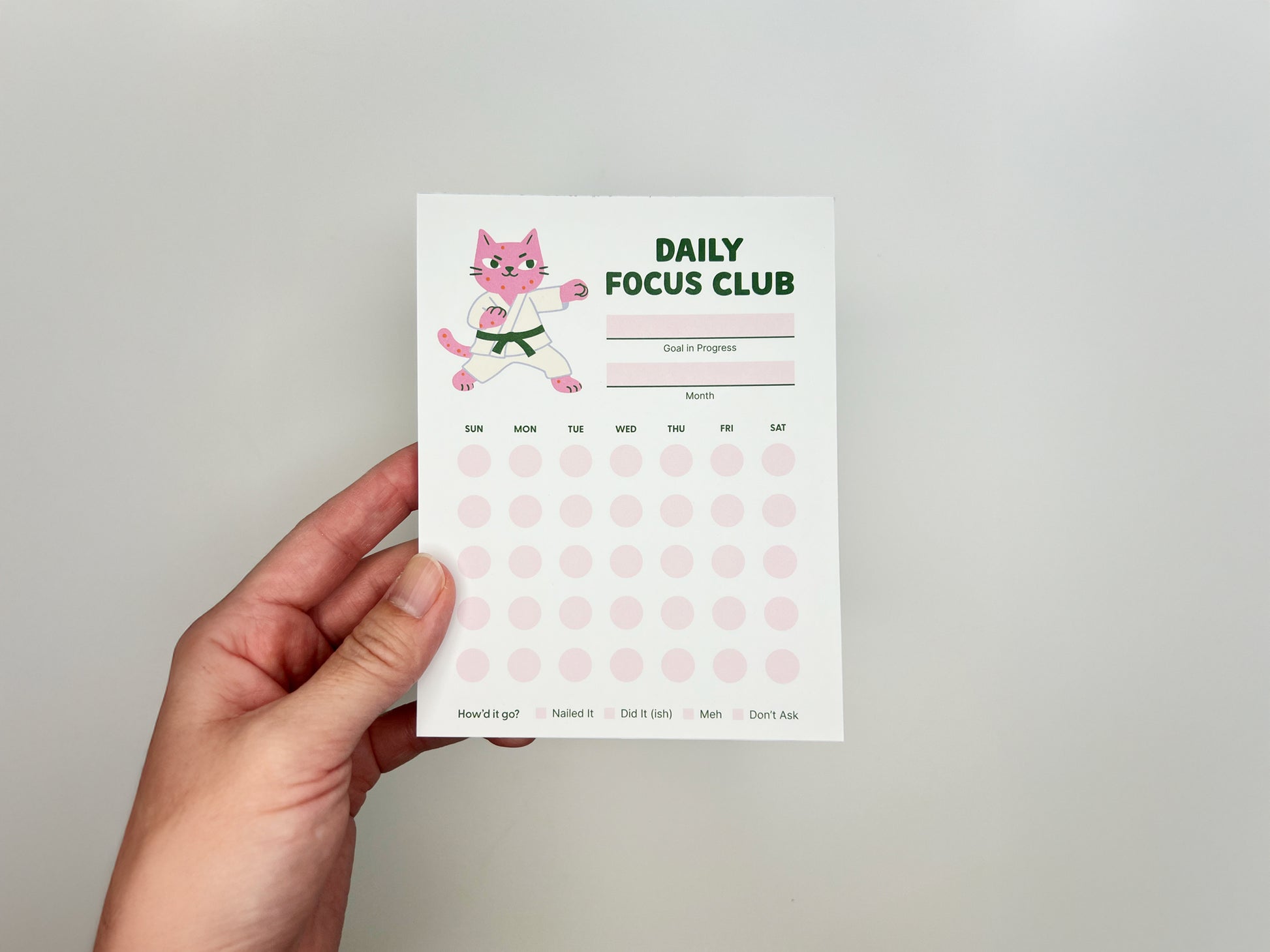 Hand holding Daily Focus Club 4.25 x 5.5 inch monthly habit tracker notepad featuring pink karate cat illustration, goal and month sections, and soft pink daily tracking dots arranged Sunday through Saturday. Cute productivity notepad for goal setting, habit tracking, ADHD-friendly planning, and desk organization by Sungy Side Up.
