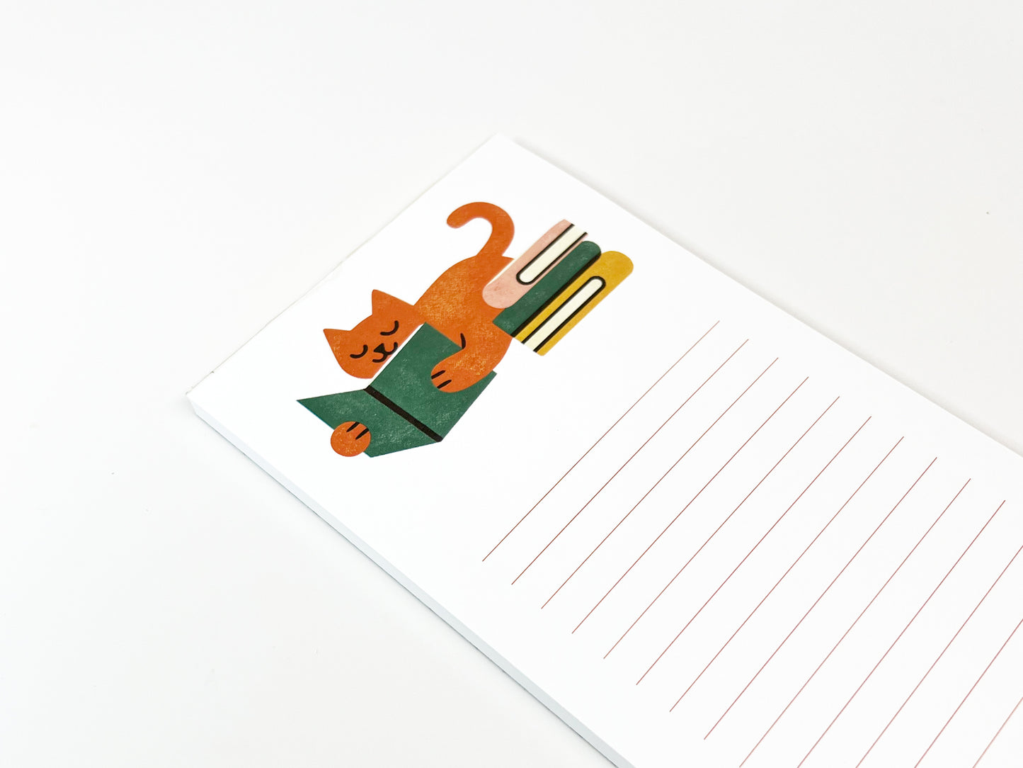 Angled close-up of a slim lined notepad showing an illustrated orange cat lounging and reading a green book beside a small stack of books, with plenty of lined writing space on a clean white background.
