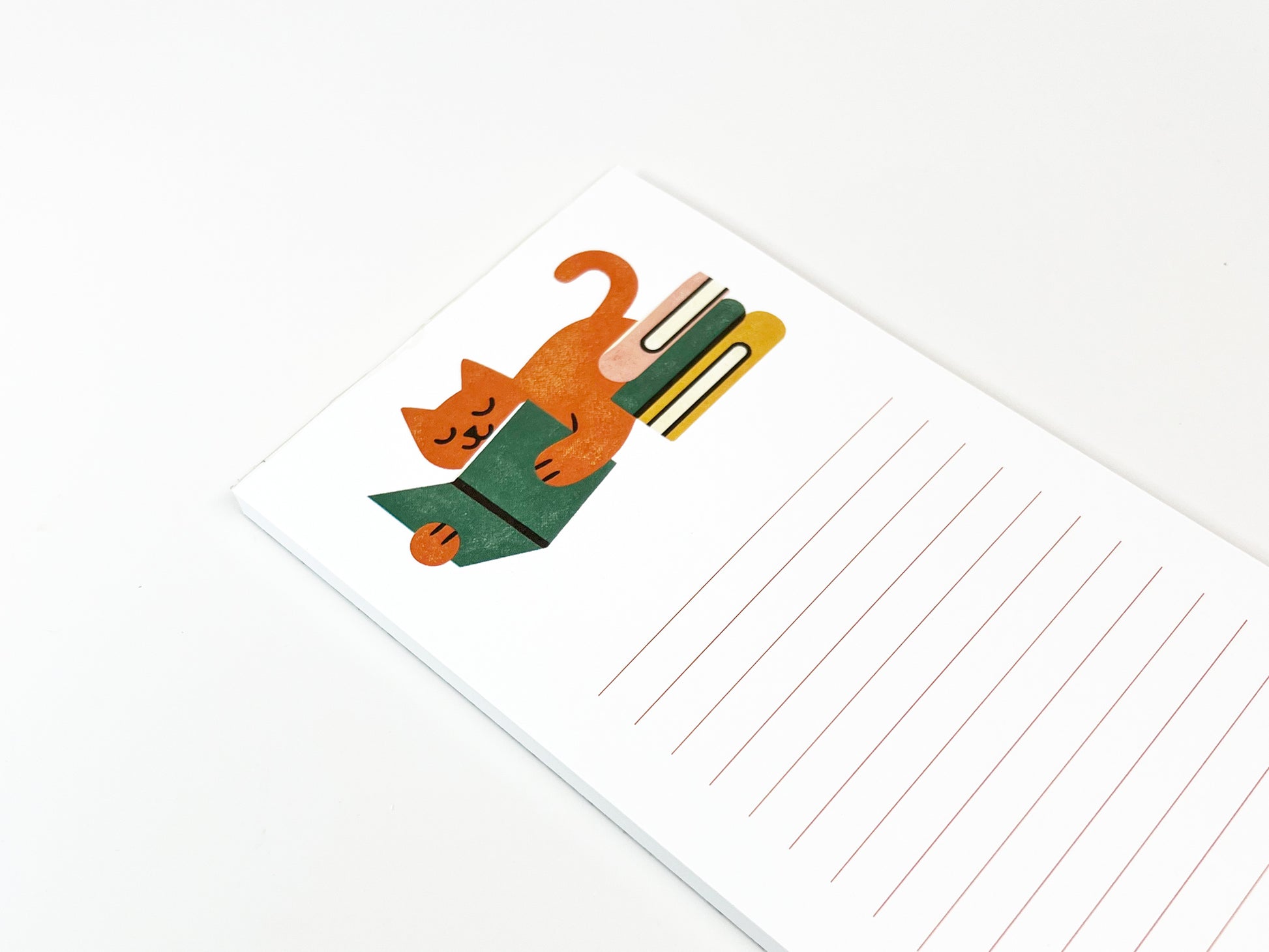 Angled close-up of a slim lined notepad showing an illustrated orange cat lounging and reading a green book beside a small stack of books, with plenty of lined writing space on a clean white background.