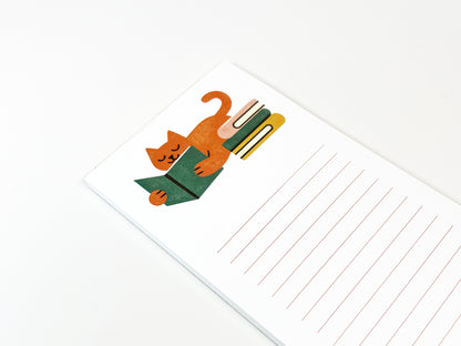 Angled close-up of a slim lined notepad showing an illustrated orange cat lounging and reading a green book beside a small stack of books, with plenty of lined writing space on a clean white background.