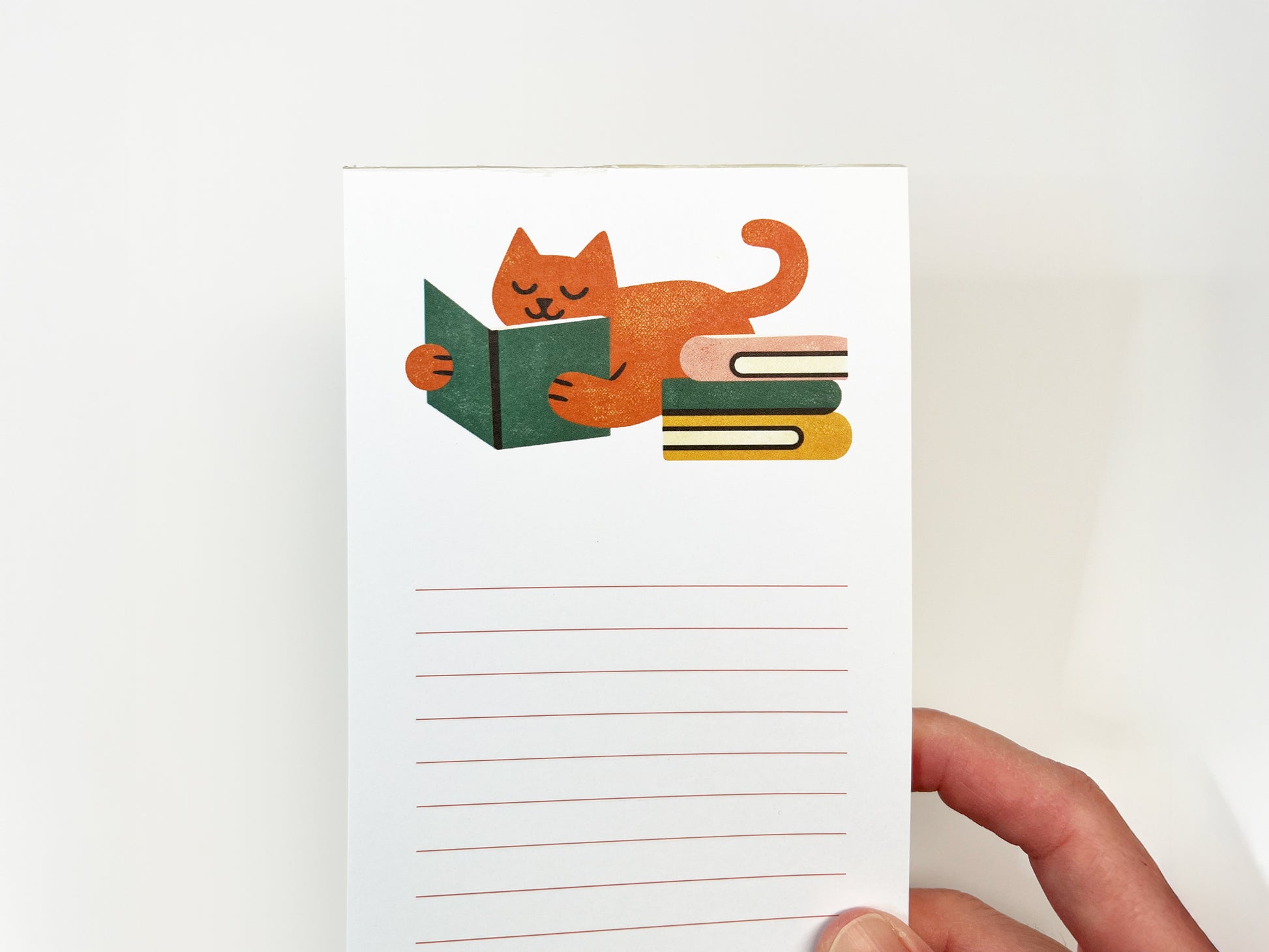 Hand holding a tall, slim lined notepad featuring an illustration of an orange cat reading a green book next to a small stack of books, with open lined space for notes on a white background.