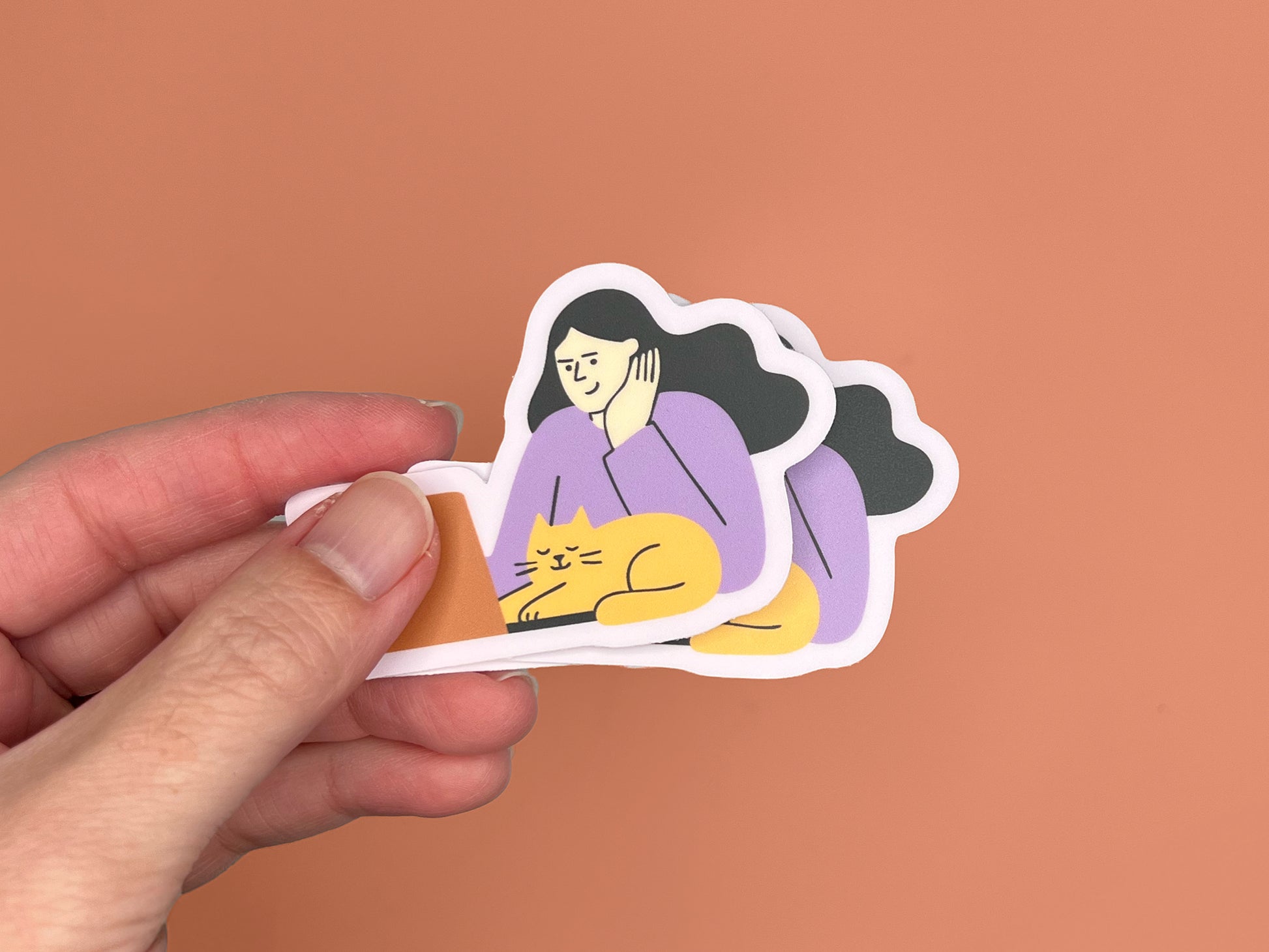A hand holding three matte vinyl die-cut stickers stacked slightly offset. Each sticker shows an illustration of a person with long dark hair wearing a lavender top, resting their head on one hand while sitting at a laptop. A sleepy yellow cat is curled in front of the laptop with eyes closed. The stickers have a soft lavender border. The background is a warm copper color.