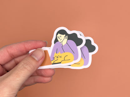 A hand holding three matte vinyl die-cut stickers stacked slightly offset. Each sticker shows an illustration of a person with long dark hair wearing a lavender top, resting their head on one hand while sitting at a laptop. A sleepy yellow cat is curled in front of the laptop with eyes closed. The stickers have a soft lavender border. The background is a warm copper color.