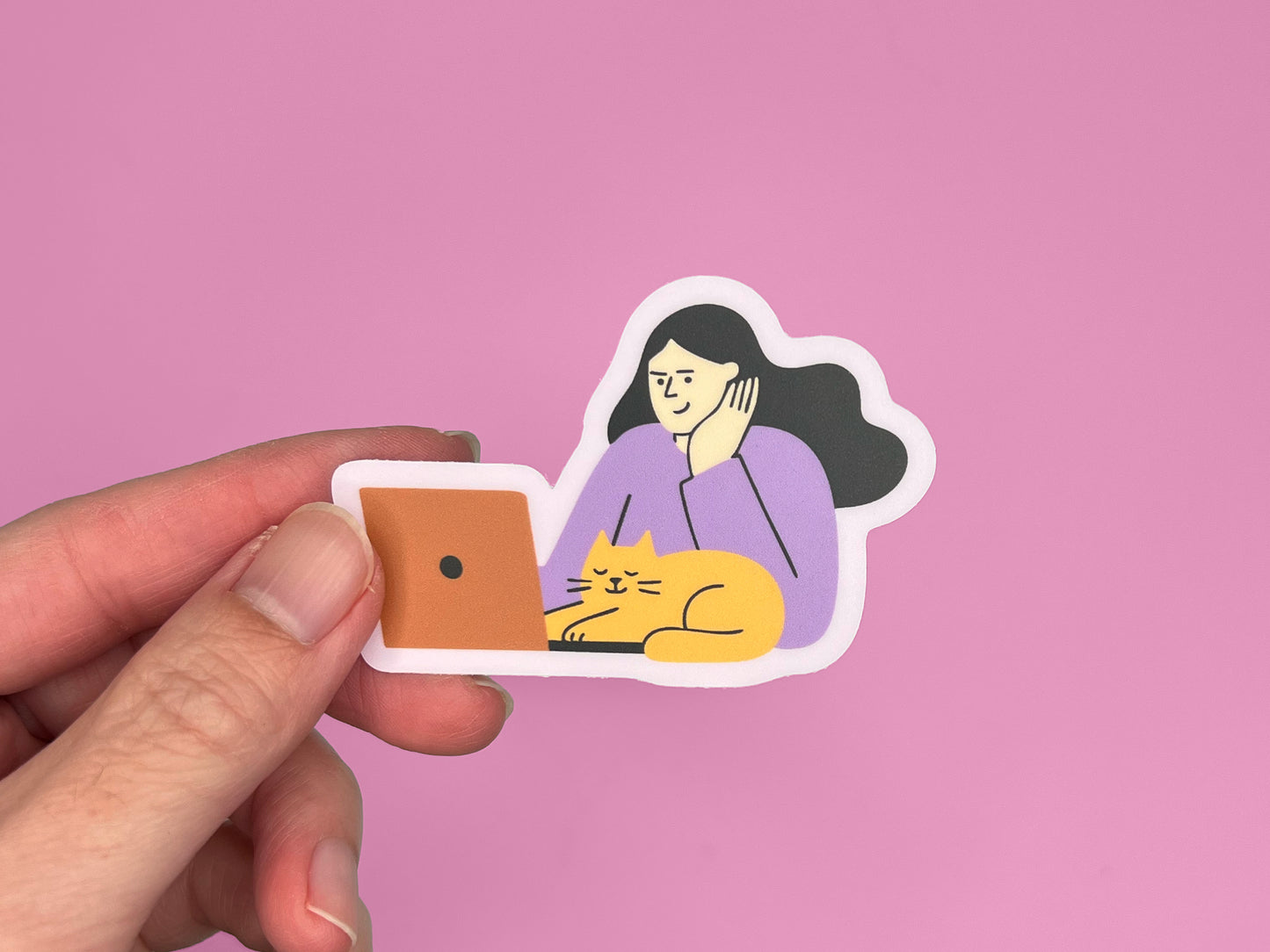 A hand holding a matte vinyl die-cut sticker against a pink background. The sticker shows an illustration of a person with long dark hair wearing a lavender top, resting their head on one hand while looking at an orange laptop. A relaxed yellow cat with closed eyes is lying in front of the laptop. The sticker has a soft lavender border around the illustration.