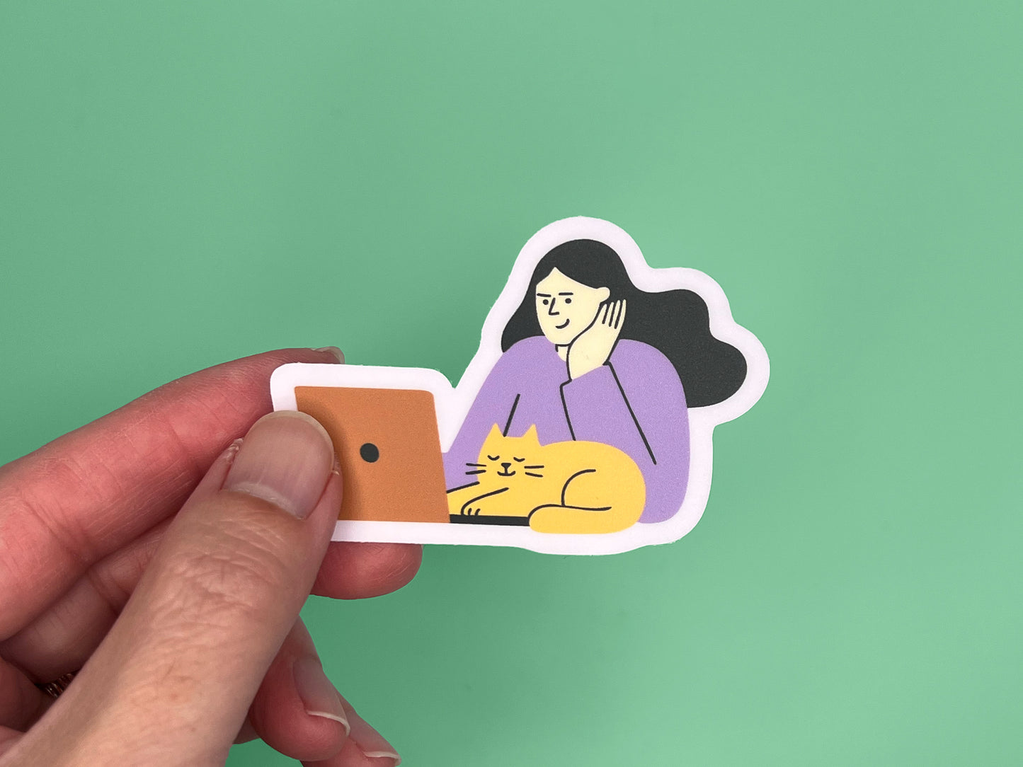 A hand holding a matte vinyl die-cut sticker against a green background. The sticker shows an illustration of a person with long dark hair wearing a lavender top, resting their head on one hand while looking at an orange laptop. A relaxed yellow cat with closed eyes is lying in front of the laptop. The sticker has a soft lavender border around the illustration.