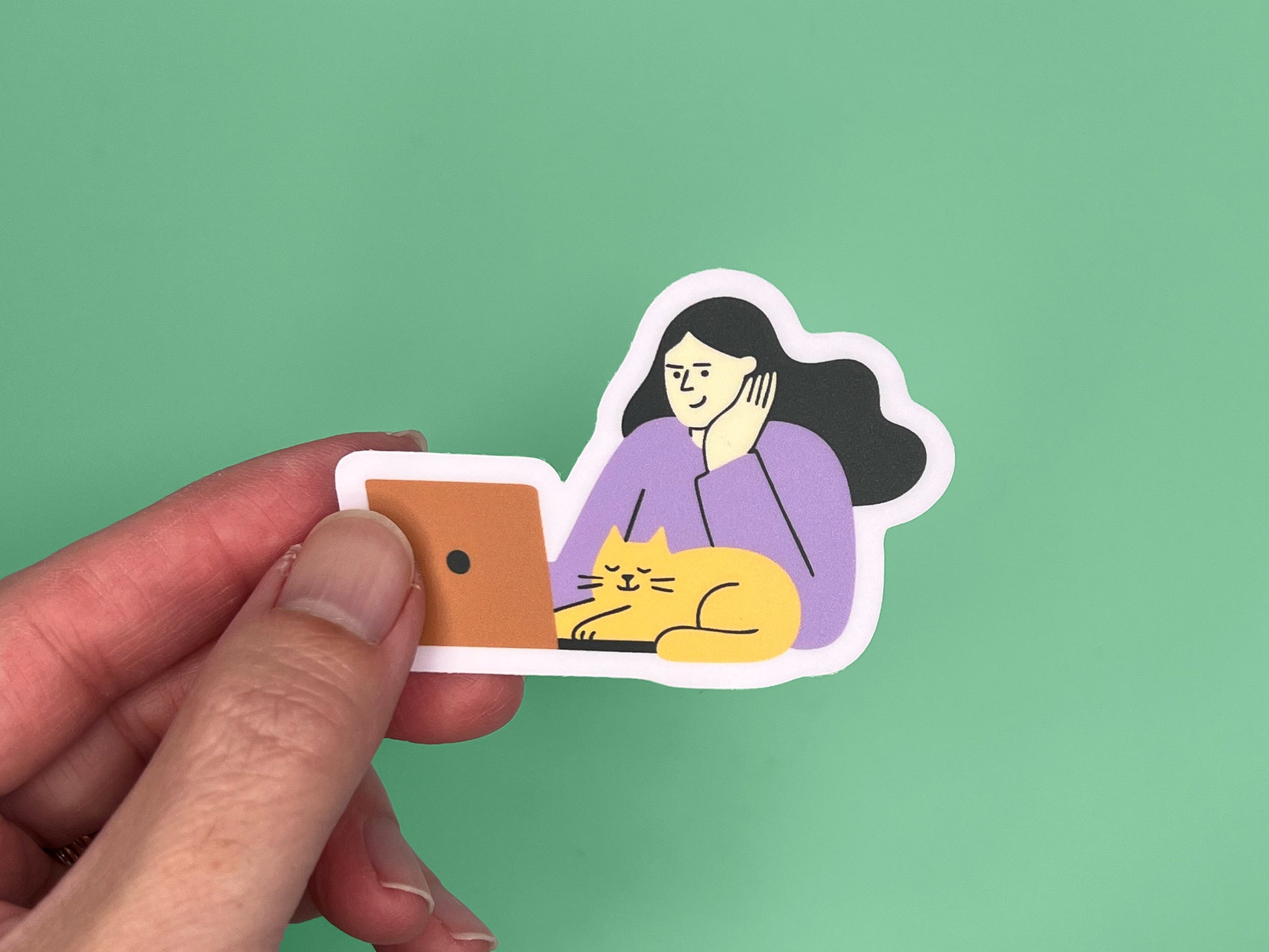 A hand holding a matte vinyl die-cut sticker against a green background. The sticker shows an illustration of a person with long dark hair wearing a lavender top, resting their head on one hand while looking at an orange laptop. A relaxed yellow cat with closed eyes is lying in front of the laptop. The sticker has a soft lavender border around the illustration.