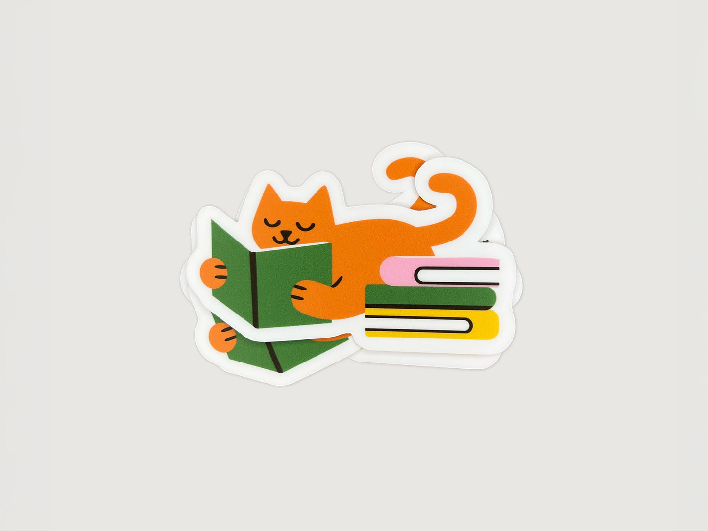 Small stack of matte vinyl die cut stickers showing an orange cat lying on its belly while reading a green book, with a curled tail and a colorful stack of books beside it. The stickers have a white border and are photographed on a light neutral background. Cute reading cat sticker for book lovers and cat lovers, made from water- and fade-resistant vinyl.