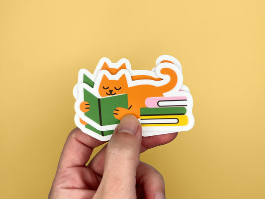 Hand holding a small stack of matte vinyl die cut stickers featuring an orange cat lying on its belly while reading a green book, with a tail curled upward and a colorful stack of books beside it. The stickers have a white border and are shown against a solid mustard yellow background. Cute reading cat sticker designed for book lovers and cat lovers, water-resistant vinyl.
