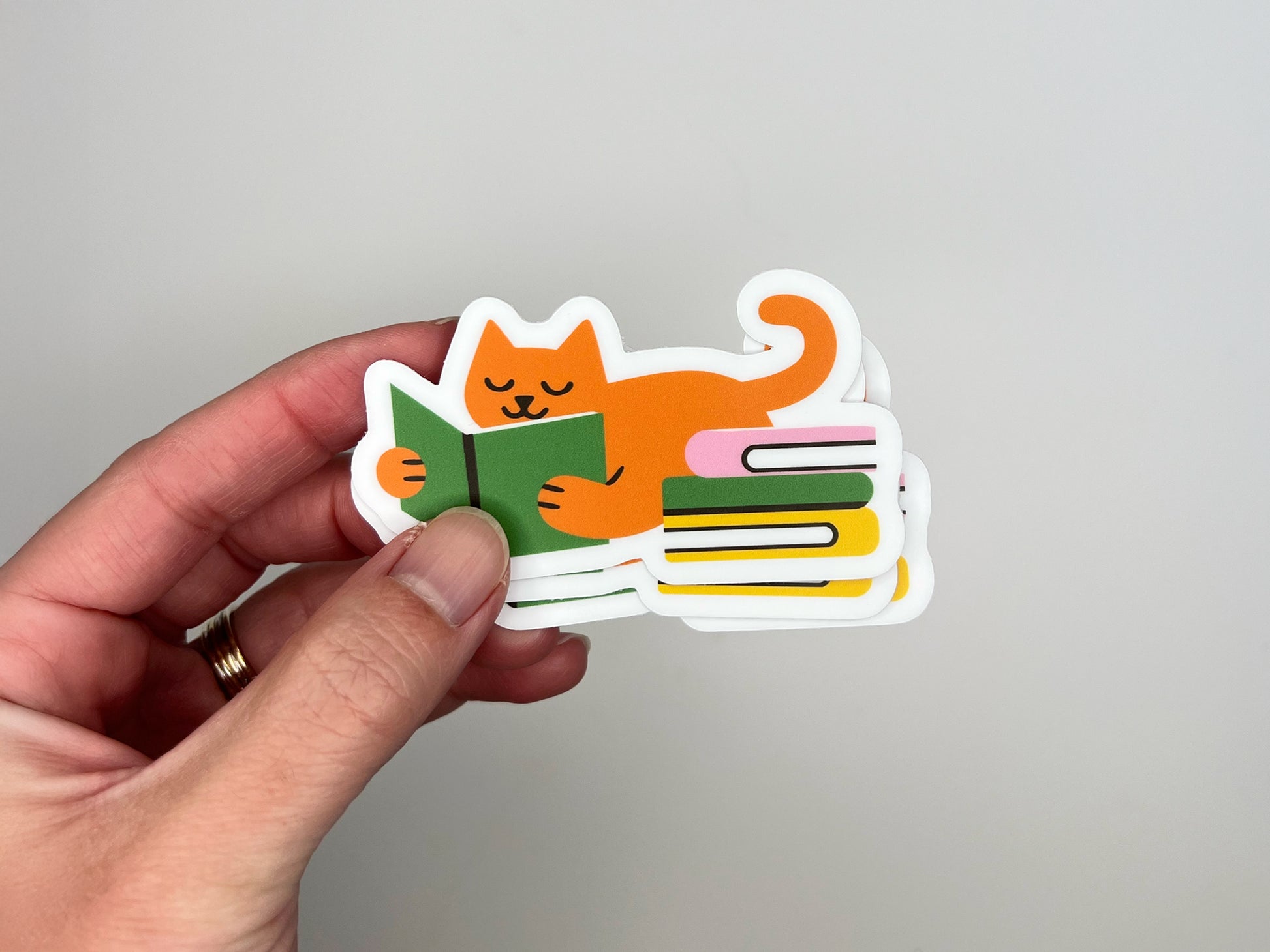 Hand holding a small stack of matte vinyl die cut stickers showing an orange cat lying on its belly while reading a green book, with its tail curled up and a colorful stack of books beside it. The stickers have a white border and are photographed against a light gray background. Cute reading cat sticker for book lovers and cat lovers, made from water-resistant vinyl.