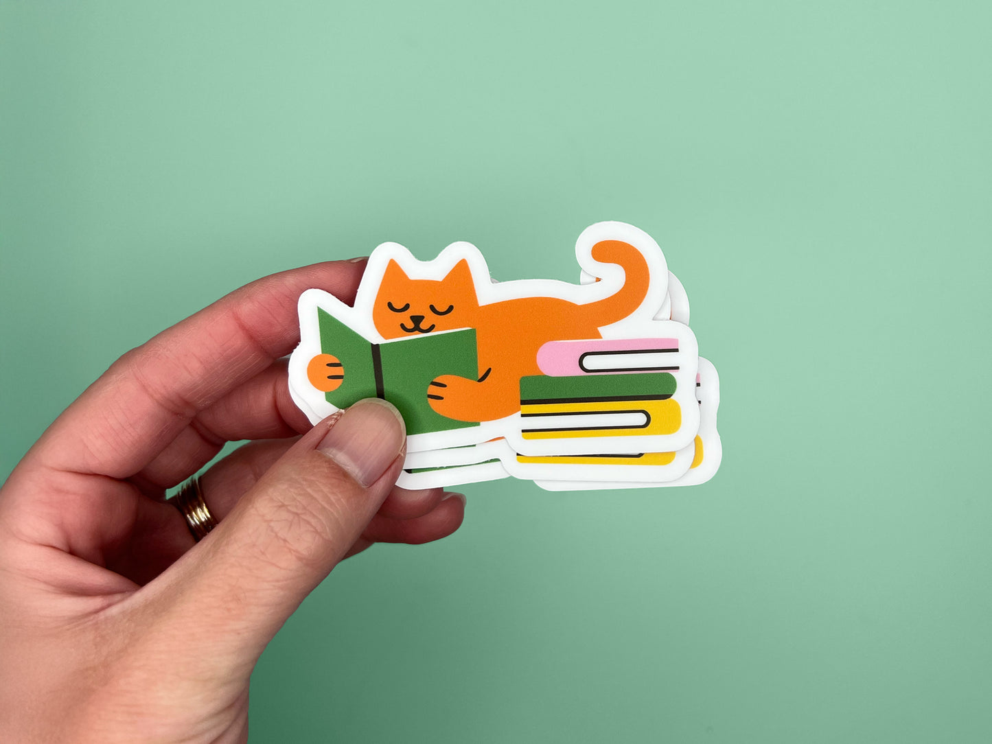 Hand holding a small stack of matte vinyl die cut stickers featuring an orange cat lying on its belly while reading a green book, with a curled tail and a colorful stack of books beside it. The stickers have a white border and are photographed against a soft green background. Cute reading cat sticker for book lovers and cat lovers, made from water- and fade-resistant vinyl.