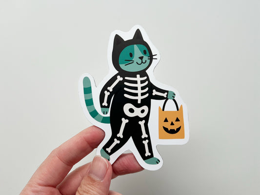 Hand holding a handmade die-cut Halloween magnet featuring a green and black cat dressed in a skeleton costume, carrying an orange pumpkin candy bag. The whimsical design is perfect for seasonal décor on fridges, lockers, or offices, appealing to cat lovers and spooky holiday enthusiasts.