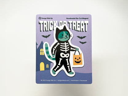 Handmade die-cut Halloween magnet featuring a green and black cat in a skeleton costume holding an orange pumpkin candy bag, displayed on a purple backing card with bats and a haunted house illustration. Perfect for seasonal décor and gifting to cat or Halloween lovers.