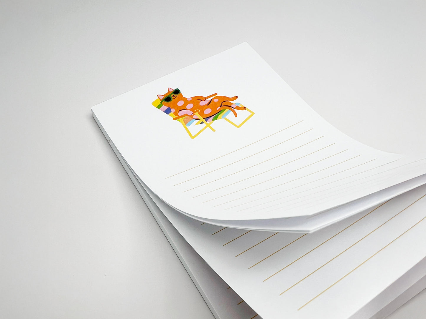 Stack of 4x6 lined notepad sheets shown at an angle on a white surface, with the top page slightly curled. The notepad features a playful illustration of a pink polka-dot cat wearing green sunglasses while relaxing on a rainbow-striped lounge chair. Light yellow writing lines appear beneath the illustration. The image highlights the thickness of the 50-sheet memo pad, designed as a cute desk notepad for cat lovers.
