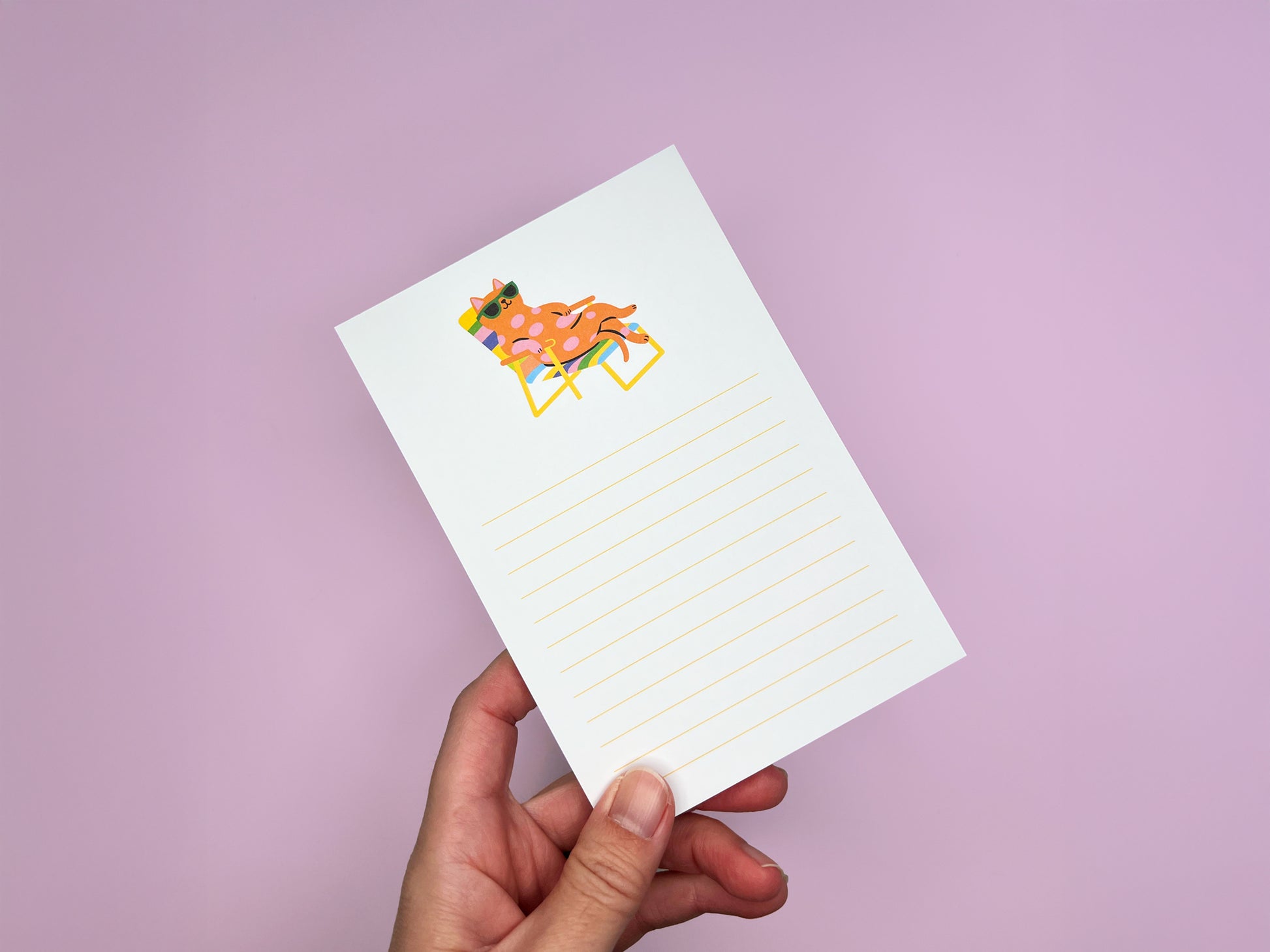Hand holding a 4x6 lined notepad against a soft pink background. The notepad features a colorful illustration at the top of a pink polka-dot cat wearing green sunglasses while lounging on a rainbow-striped beach chair. Light yellow writing lines fill the lower portion of the page. The image shows a playful, minimalist memo pad designed for cat lovers, perfect for desk notes, reminders, or gifting.