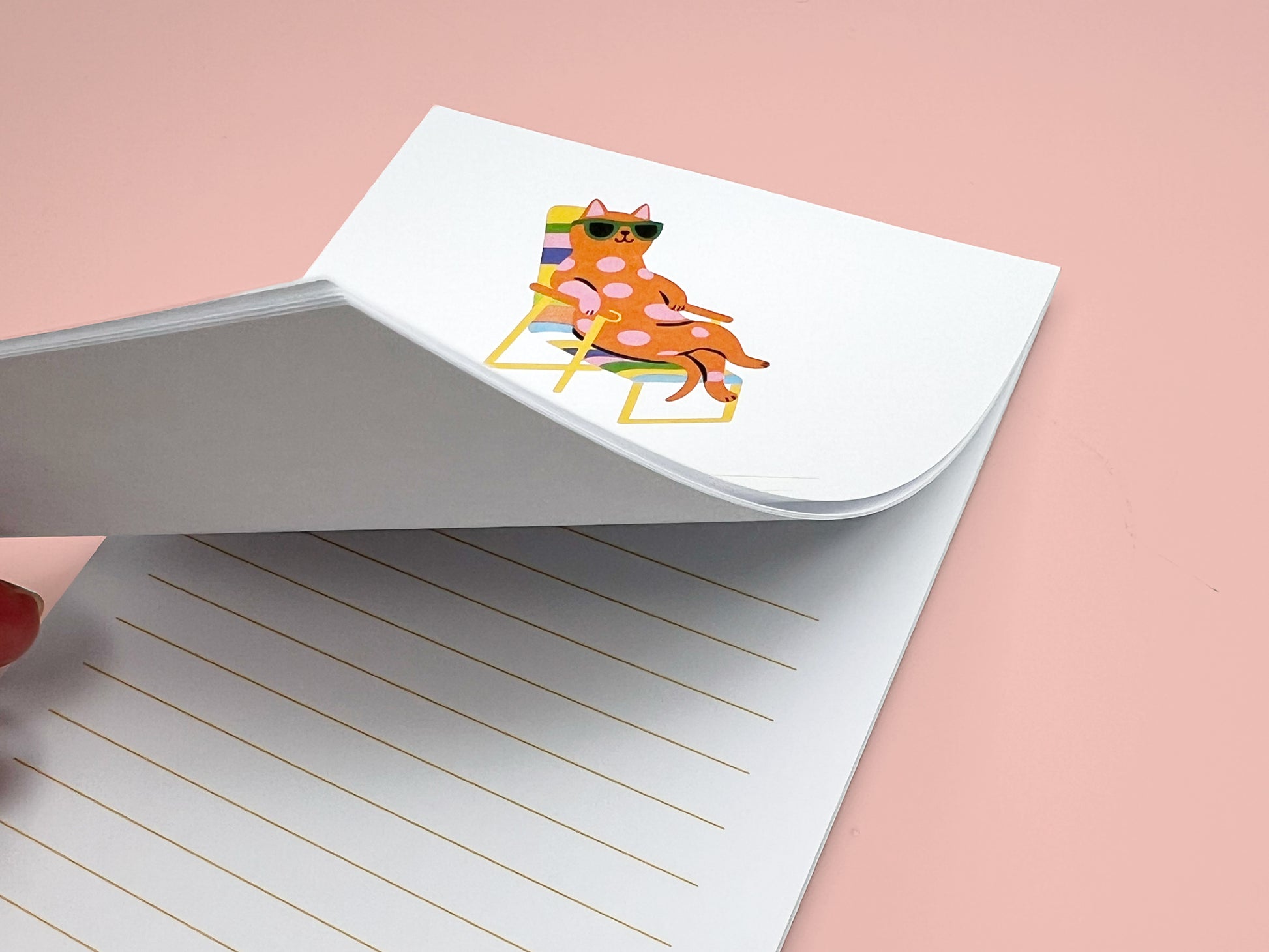 Close-up of a 4x6 lined notepad with the top sheet partially lifted to show the thickness of the paper stack. The notepad features a playful illustration of a pink polka-dot cat wearing green sunglasses while lounging on a rainbow-striped beach chair. Light yellow writing lines appear on each page. The image highlights the quality and thickness of the 50-sheet memo pad, perfect for desk notes, reminders, or gifts for cat lovers.