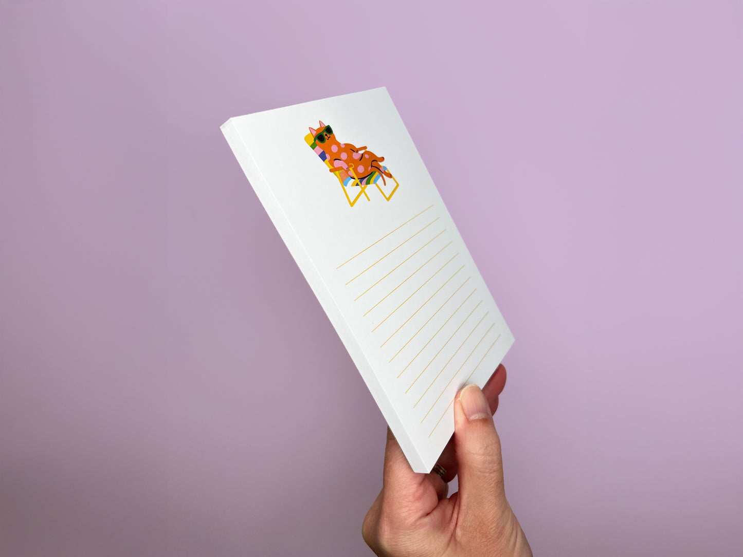 Hand holding a 4x6 lined notepad at an angle against a soft lavender background, showing the thickness of the 50-sheet memo pad. The top page features a colorful illustration of a pink polka-dot cat wearing green sunglasses while lounging on a rainbow-striped beach chair. Light yellow writing lines appear on each sheet, highlighting the playful design and sturdy paper quality of this cute desk notepad for cat lovers.
