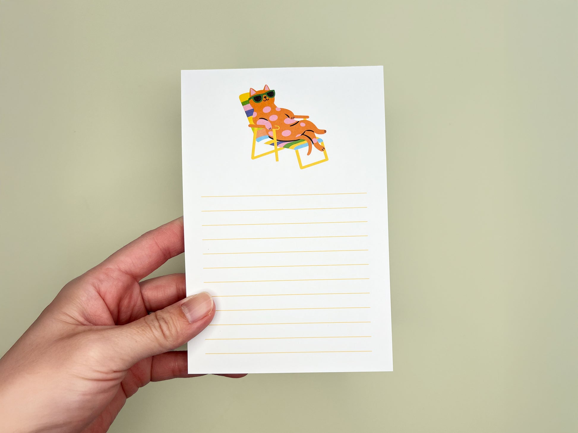 Hand holding a 4x6 lined notepad against a soft pale green background. The notepad features a colorful illustration at the top of a pink polka-dot cat wearing green sunglasses while lounging back in a rainbow-striped beach chair. Below the illustration are light yellow horizontal writing lines across the page. The notepad appears clean and minimal, with a playful, relaxed summer vibe, designed as a cute memo pad for cat lovers.