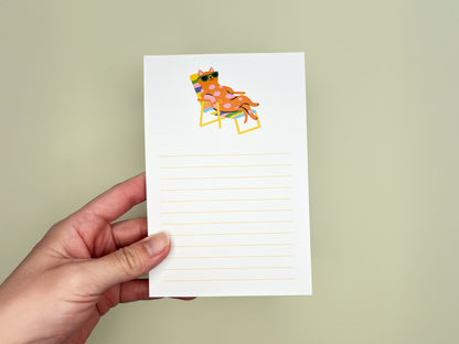 Hand holding a 4x6 lined notepad against a soft pale green background. The notepad features a colorful illustration at the top of a pink polka-dot cat wearing green sunglasses while lounging back in a rainbow-striped beach chair. Below the illustration are light yellow horizontal writing lines across the page. The notepad appears clean and minimal, with a playful, relaxed summer vibe, designed as a cute memo pad for cat lovers.