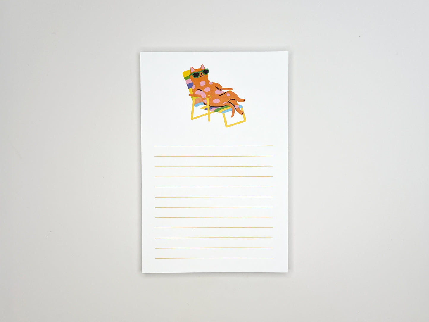 Flat lay of a 4x6 lined notepad centered on a white background, featuring a colorful illustration at the top of a pink polka-dot cat wearing green sunglasses while relaxing on a rainbow-striped lounge chair. Light yellow writing lines fill the lower half of the page. The minimalist memo pad has a playful summer vibe and is designed as a cute desk notepad for cat lovers, perfect for notes, reminders, or gifting.