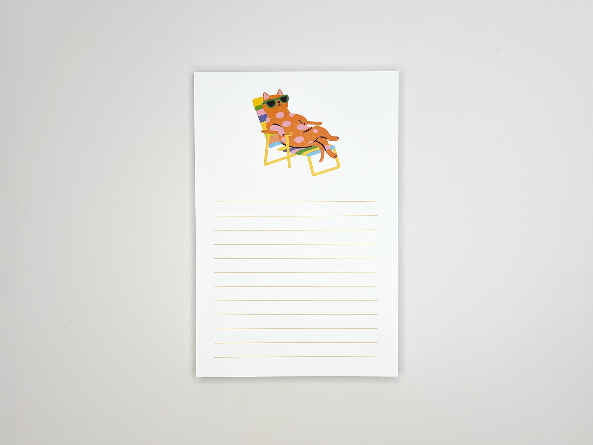 Flat lay of a 4x6 lined notepad centered on a white background, featuring a colorful illustration at the top of a pink polka-dot cat wearing green sunglasses while relaxing on a rainbow-striped lounge chair. Light yellow writing lines fill the lower half of the page. The minimalist memo pad has a playful summer vibe and is designed as a cute desk notepad for cat lovers, perfect for notes, reminders, or gifting.