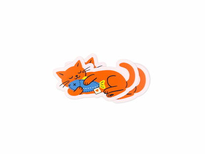 Stack of matte vinyl die cut stickers featuring a bright orange cat illustration lying on its side while hugging a blue fish toy. Cute waterproof cat stickers for laptops, water bottles, planners, and notebooks on a white background.