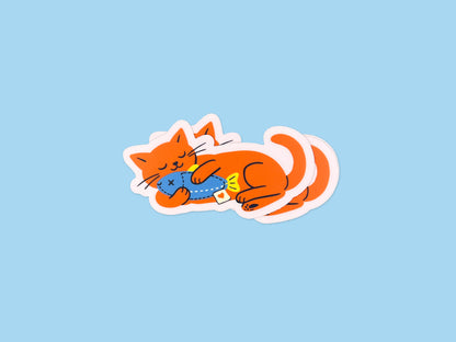 Stack of matte vinyl die cut stickers showing a bright orange cat illustration lying on its side while hugging a blue fish toy. Cute waterproof cat stickers on a light blue background for laptops, water bottles, planners, and notebooks.