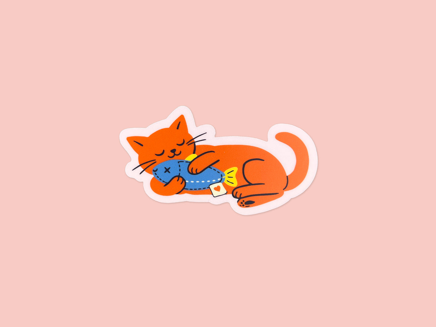 Single matte vinyl die cut sticker of a bright orange cat illustration lying on its side while hugging a blue fish toy. Cute waterproof cat sticker on a soft pink background for laptops, water bottles, planners, and notebooks.