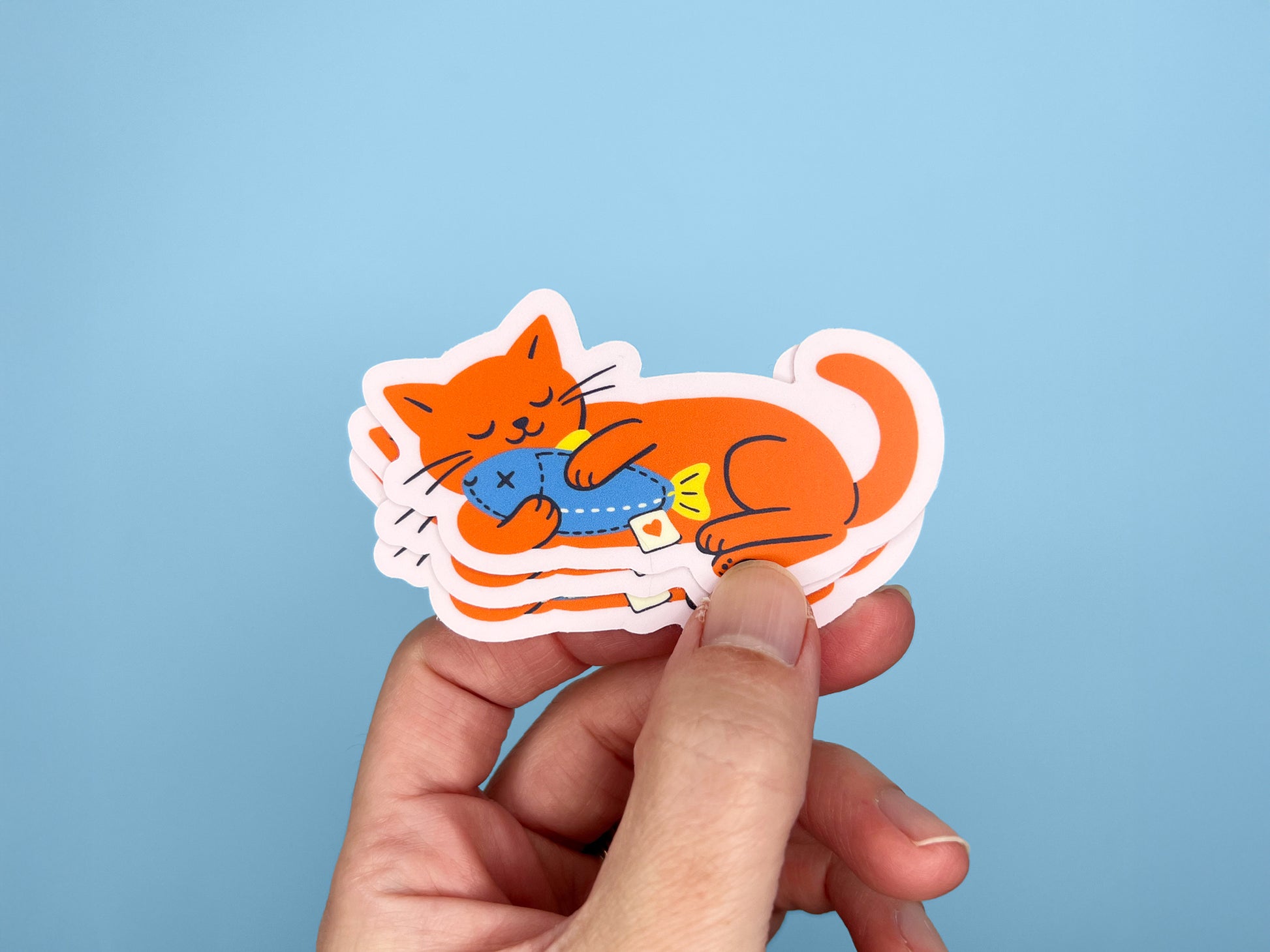 Hand holding a small stack of matte vinyl die cut stickers featuring a bright orange cat illustration lying down and hugging a blue fish toy. Cute waterproof cat stickers for laptops, water bottles, planners, and notebooks on a light blue background.
