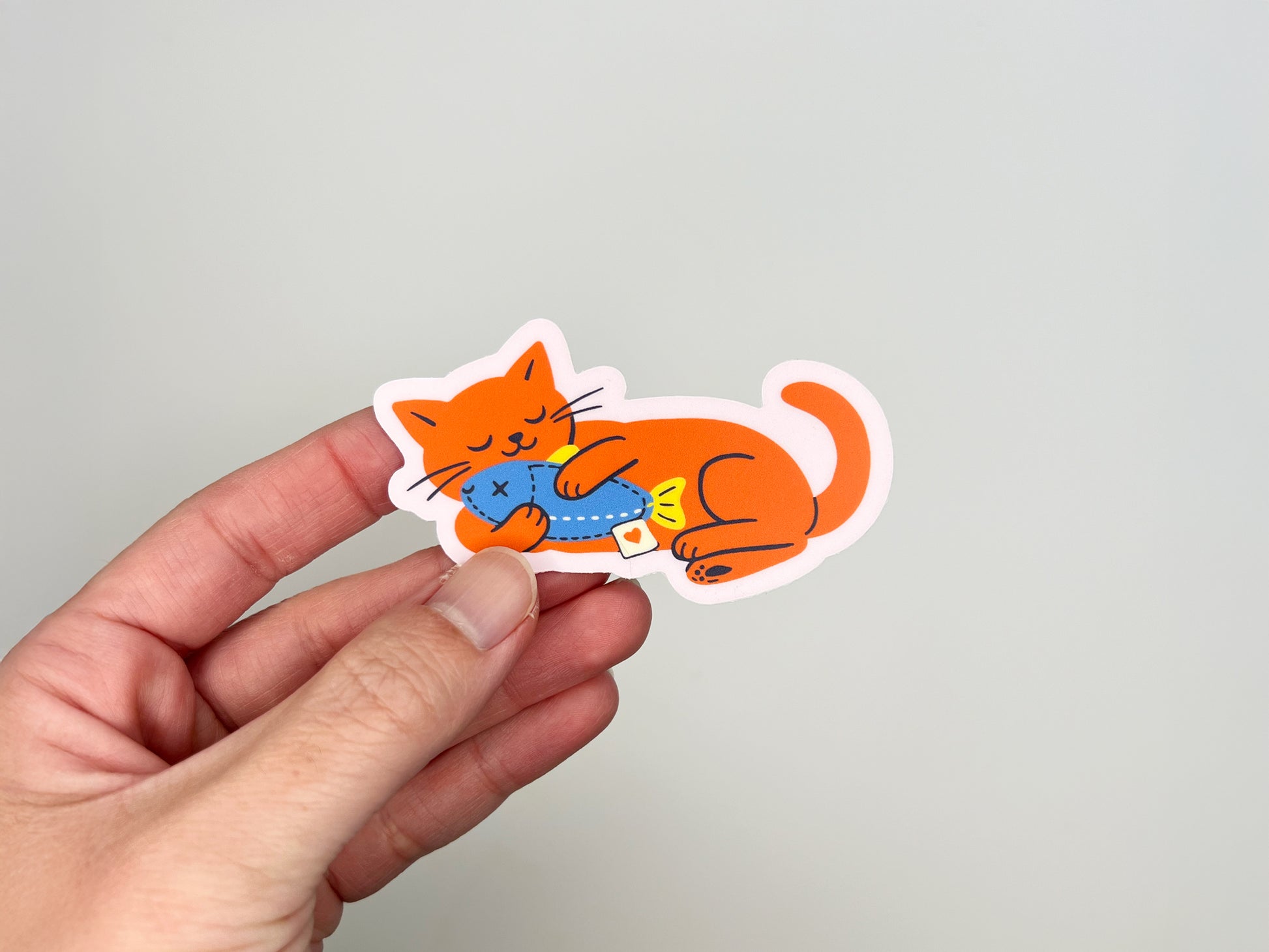 Hand holding a matte vinyl die cut sticker of a bright orange cat illustration lying on its side while hugging a blue fish toy. Cute waterproof cat sticker for laptops, water bottles, planners, and notebooks on a neutral background.