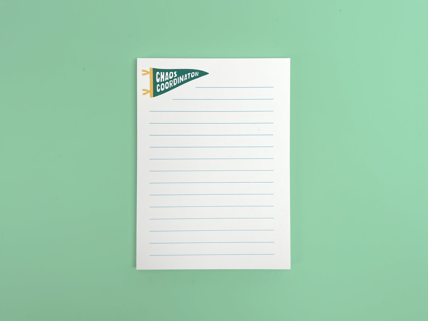 Alt text: White lined notepad with a small green pennant flag illustration reading “Chaos Coordinator” in bold white lettering at the top left corner. The notepad is photographed flat against a mint green background and features light blue horizontal writing lines with a clean, minimal design.