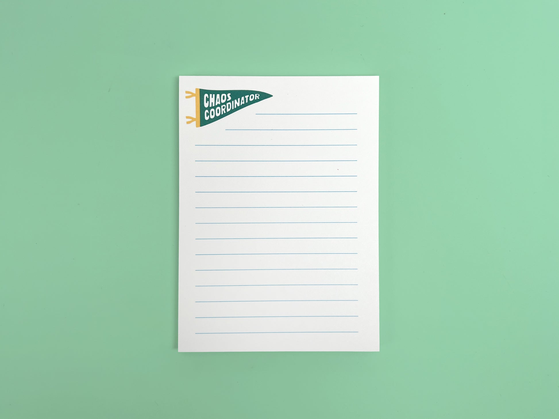 Alt text: White lined notepad with a small green pennant flag illustration reading “Chaos Coordinator” in bold white lettering at the top left corner. The notepad is photographed flat against a mint green background and features light blue horizontal writing lines with a clean, minimal design.
