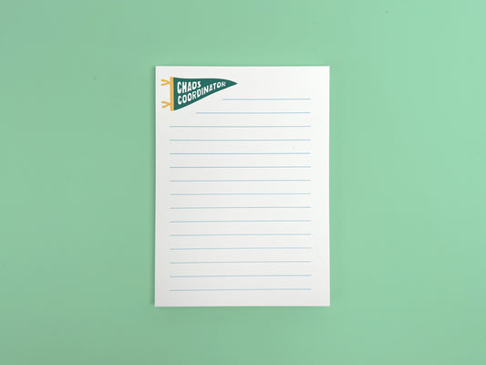 Alt text: White lined notepad with a small green pennant flag illustration reading “Chaos Coordinator” in bold white lettering at the top left corner. The notepad is photographed flat against a mint green background and features light blue horizontal writing lines with a clean, minimal design.