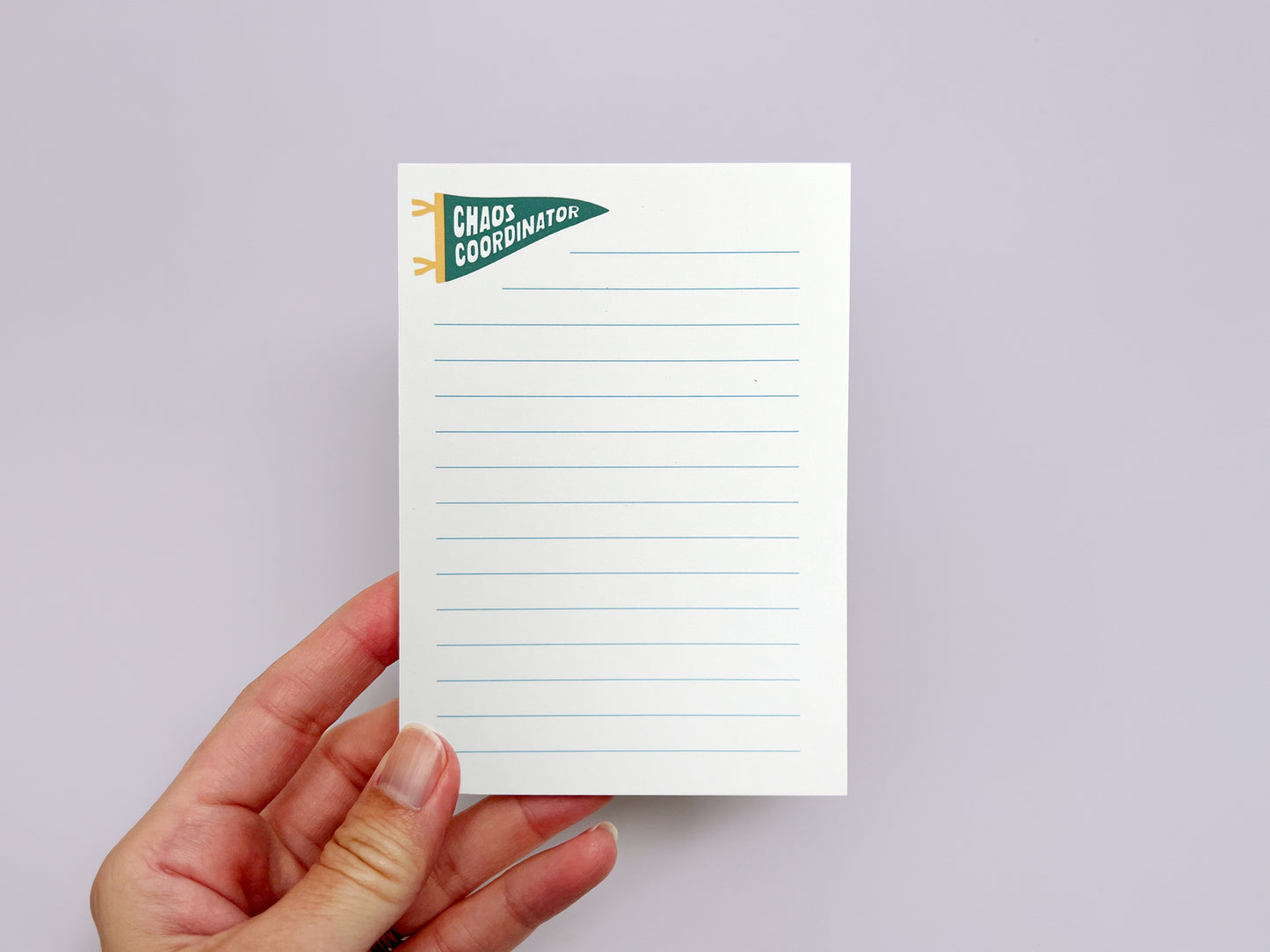 Alt text: Hand holding a white lined notepad with a green pennant illustration that reads “Chaos Coordinator” in bold white text near the top corner. The notepad features pale blue writing lines and is photographed against a soft light lavender background.
