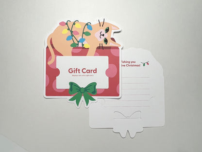 A close-up of the holiday gift card holder with a festive cat lounging on a present. The holder has a space on the front for the gift card and a green bow for decoration. The back is visible, showing room for a personal message with the words "Wishing you a Festive Christmas!" and a small string of holiday lights.