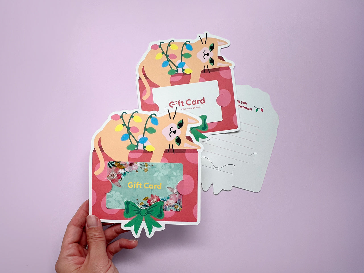 A cute and festive Christmas cat gift card holder featuring a playful yellow cat tangled in colorful holiday lights, sitting on a red and pink gift box with a green bow. This whimsical design makes it the perfect for cat lovers.