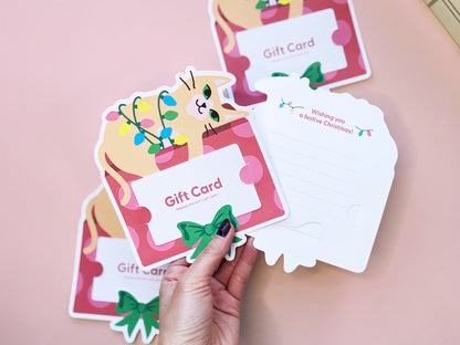 Hand holing a gift card holder to showcase a design of an orange cat wrapped in colorful holiday lights sitting on a red and pink gift box with a festive green bow, complete with slots for inserting a gift card. The back has space a personal note.