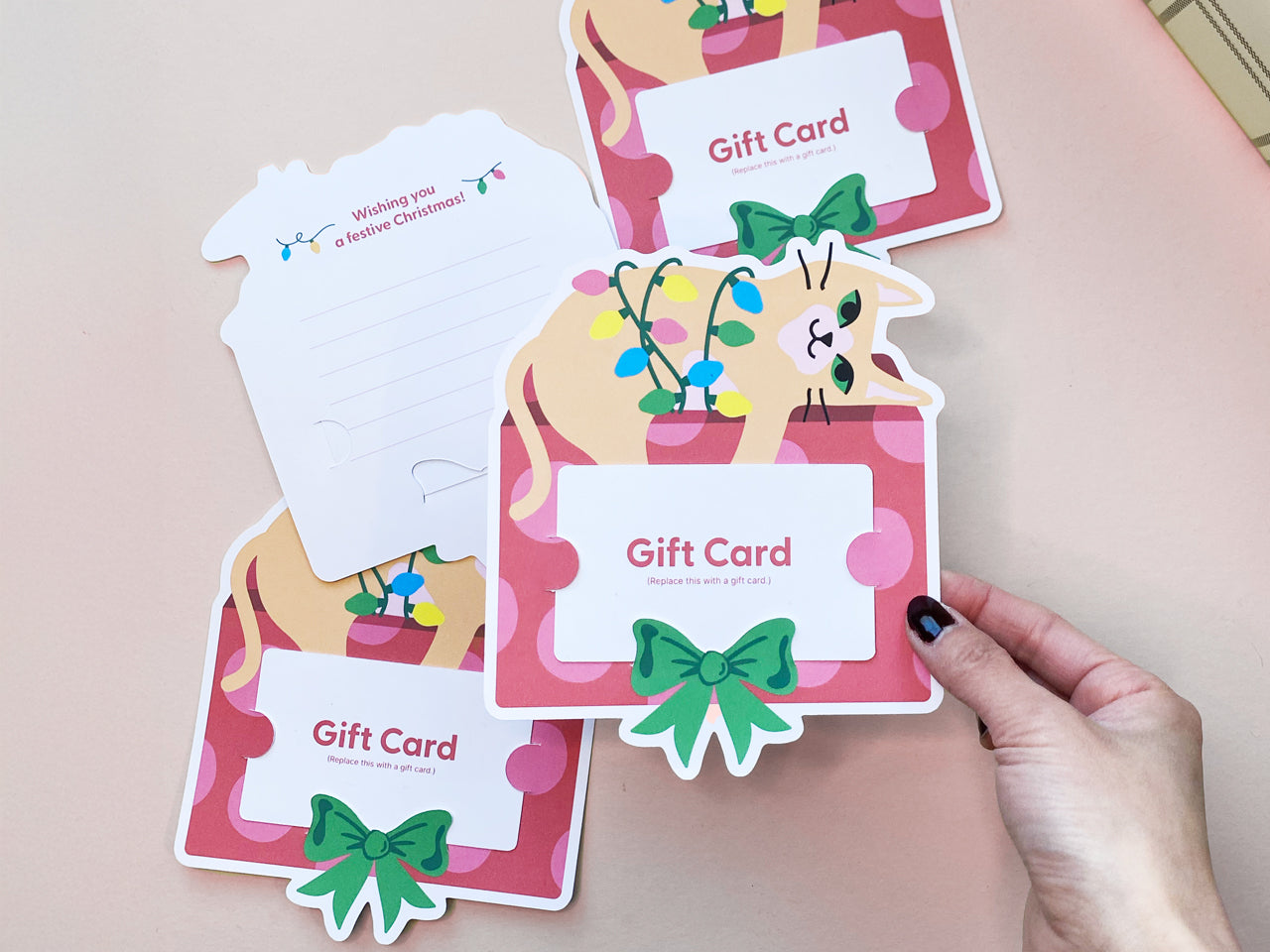 Hand holing a gift card holder to showcase a design of an orange cat wrapped in colorful holiday lights sitting on a red and pink gift box with a festive green bow, complete with slots for inserting a gift card. The back has space a personal note.