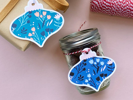 Two handmade die-cut Christmas ornament-shaped gift tags with floral patterns in teal and blue. One tag is tied to a mason jar with red and white twine, while the other is attached to a wrapped gift with a mustard-colored ribbon.
