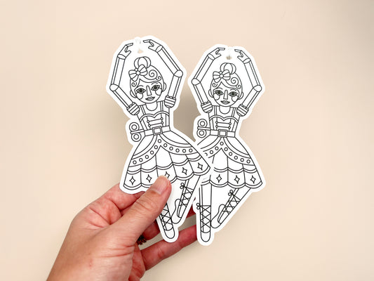 A hand holding two identical nutcracker ballerina-shaped gift tags with black-and-white line art designs. Each tag shows a ballerina wearing a bow and tutu with raised arms, printed on white cardstock, displayed against a soft beige background.