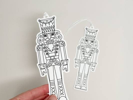 A hand holding one of two nutcracker-shaped gift tags with black-and-white line art designs. Both tags feature the same nutcracker illustration with white string attached, displayed against an off-white background.