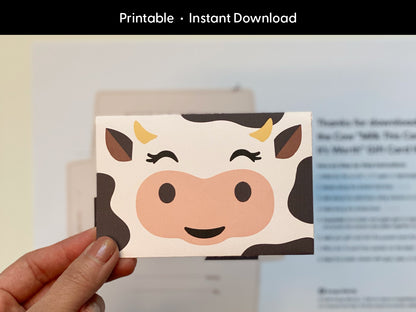 Hand holding an assembled printable cow gift card holder featuring a cute cartoon cow face, with blurred instructions in the background. This digital download template is perfect for gifting teachers, kids, or friends during birthdays, baby showers, or holidays. Designed for easy at-home crafting, this DIY cow envelope adds a fun farm animal twist to personalized gift card giving. Instant download for quick use.