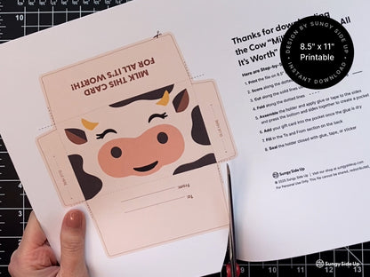 Hand holding a printed 8.5 by 11 inch sheet featuring a cow gift card holder template and assembly instructions. The cutout design includes a cute cow face and To and From section. This printable envelope is a digital download for crafting unique gift card holders at home. Perfect for birthdays, teacher appreciation, baby showers, and party favors. Designed for budget-friendly DIY gifting with a farm animal theme.