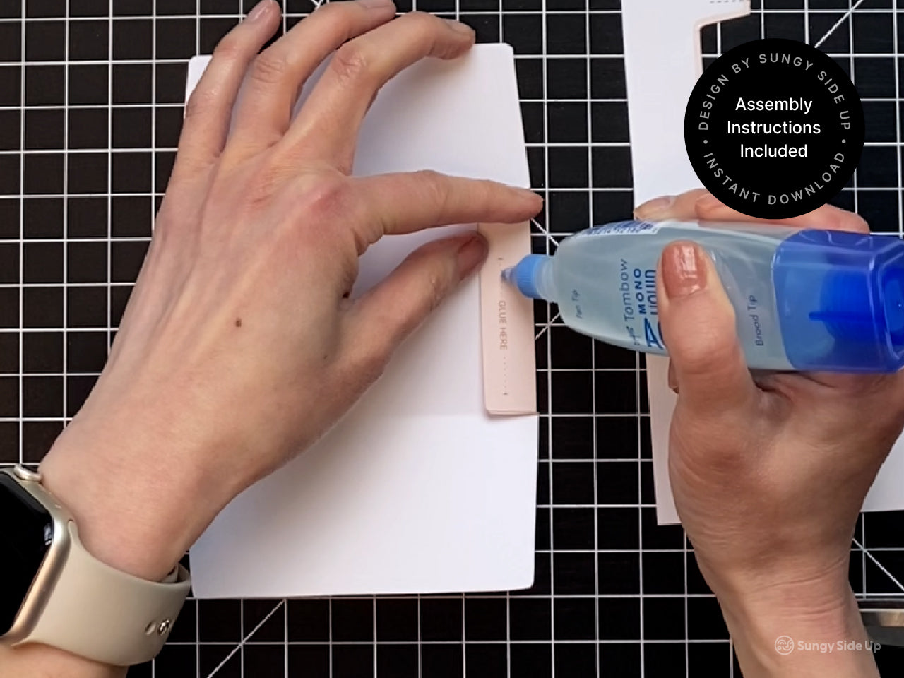 Close-up of hands applying glue to a printable cow-themed gift card holder template using a blue glue bottle. The template is laid on a black grid cutting mat, showing the easy DIY assembly process. This instant download includes full instructions and is designed for quick at-home crafting. Great for gifting teachers, kids, or party guests, this printable gift card envelope adds a fun farm animal twist to your gift.