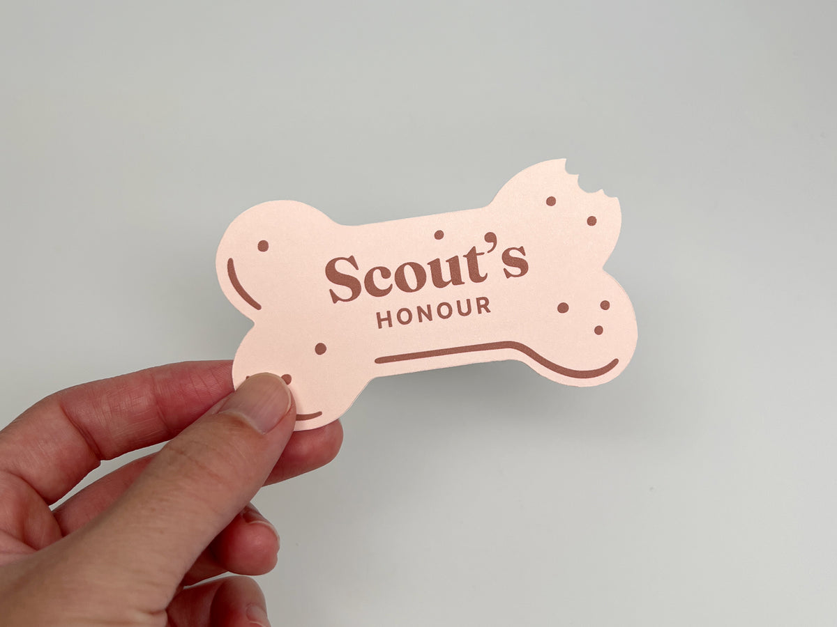 Hand holding a pink, bone-shaped dog bakery business card with a bitten corner and “Scout’s Honour” printed on the front.