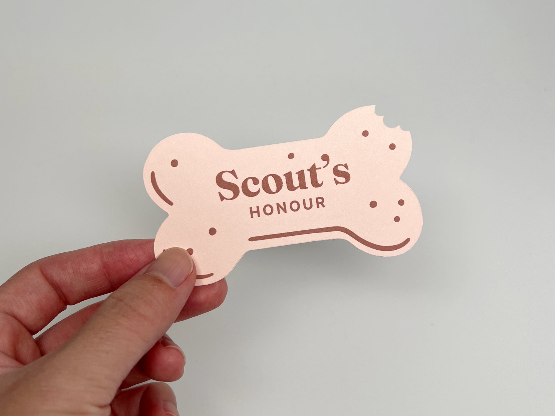 Hand holding a pink, bone-shaped dog bakery business card with a bitten corner and “Scout’s Honour” printed on the front.